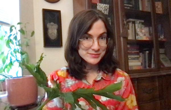 Annette LePique smiles with a leafy houseplant in her lap while sitting in front of a bookcase to the right of a window